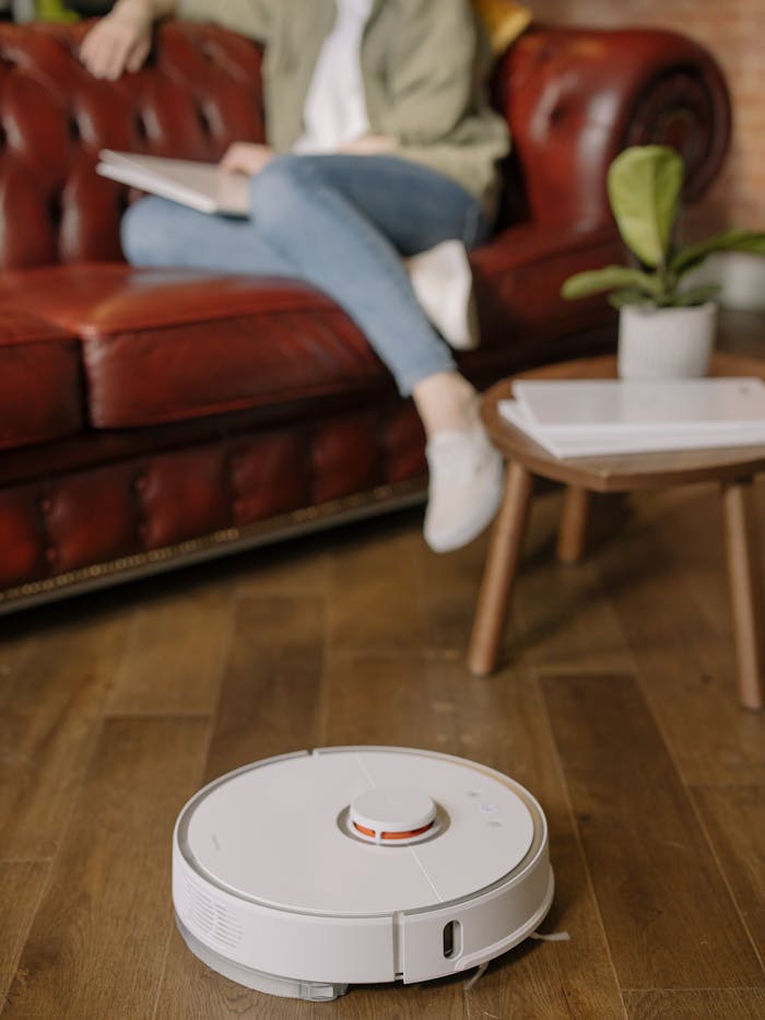 A woman relaxes on a leather sofa as a robotic vacuum cleans the hardwood floor in a cozy home.